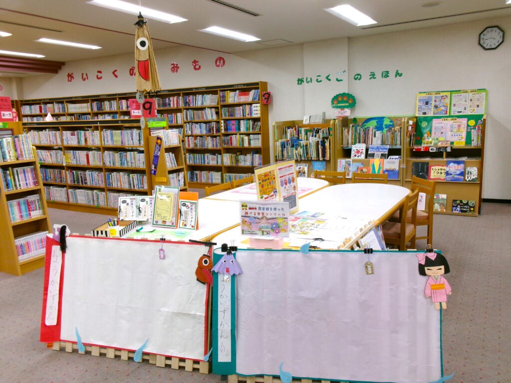 コーナー画像。唐笠お化けが吊り下がる下に大きな巻物「ようかいずかん」、机の上にみんなで書く「ようかい図鑑」の用紙、鉛筆など。