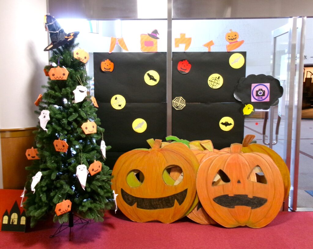 ハロウィンフォトコーナーの画像。黒い背景の前に、顔出しできるかぼちゃのパネルと、ハロウィンの飾り付けがしてあるハロウィンツリーがある。