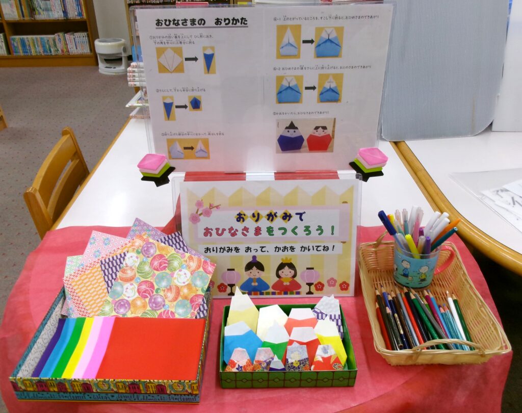 「おりがみで おひなさまを つくろう！」コーナーの画像。和柄や赤色、青色、黄色などの折り紙。色鉛筆、カラーペン。顔を描けば完成する折り紙のおひなさまが置いてある。